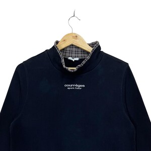 May include: Black long-sleeve sweatshirt with a high collar and a plaid pattern on the inside. The brand name "courr&egrave;ges sport futur" is printed in white on the front. The sweatshirt is hanging on a wooden hanger.