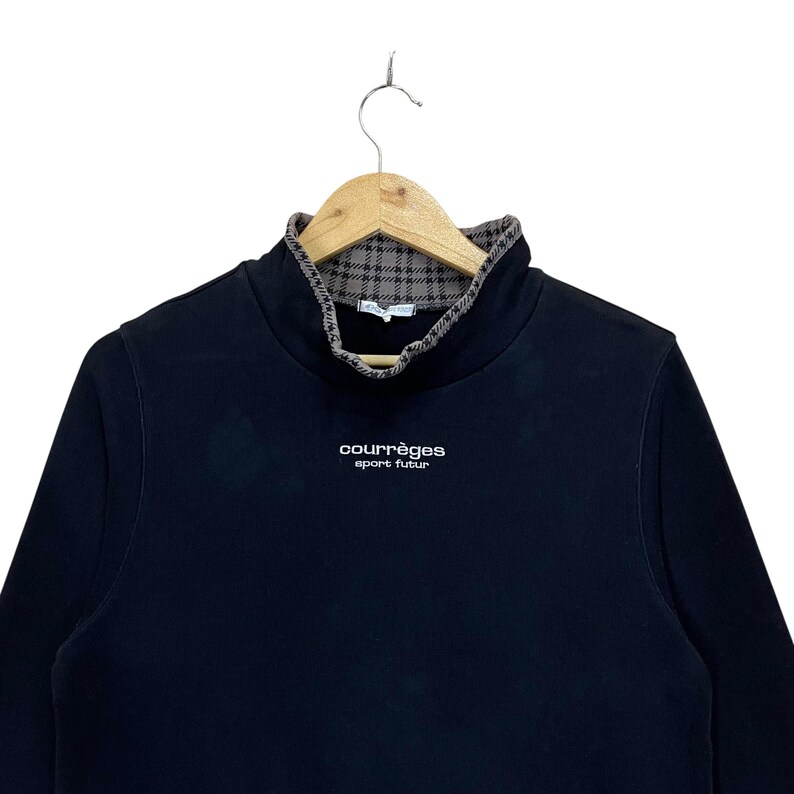 May include: Black long-sleeve sweatshirt with a high collar and a plaid pattern on the inside. The brand name "courr&egrave;ges sport futur" is printed in white on the front. The sweatshirt is hanging on a wooden hanger.