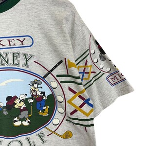 Puede incluir: Una camiseta gris con cuello verde con un gr&aacute;fico colorido de Mickey Mouse y Goofy jugando al golf. La camiseta tiene las palabras "Disney Golf" y "MICKEY" impresas.