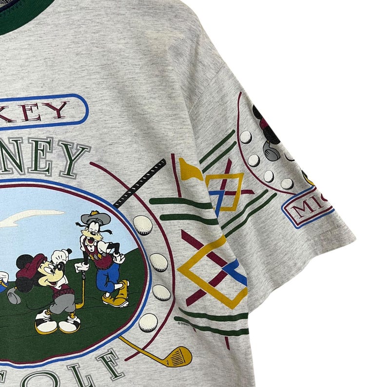 Puede incluir: Una camiseta gris con cuello verde con un gr&aacute;fico colorido de Mickey Mouse y Goofy jugando al golf. La camiseta tiene las palabras "Disney Golf" y "MICKEY" impresas.