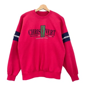 Puede incluir: Una sudadera roja con el texto "CHRIS EVERT" en negro y verde. Las mangas tienen rayas azul marino con texto blanco. La sudadera incluye las frases "THIS IS A THEMEAN FOR" y "REFRESHING MIND OF A COURT".
