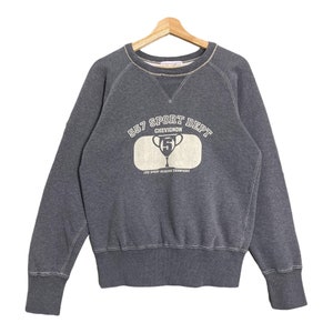 Puede incluir: Una sudadera gris con el texto "557 SPORT DEPT CHEVIGNON" en blanco y un gráfico de trofeo. La sudadera tiene cuello redondo, mangas largas y puños y dobladillo acanalados. La prenda se muestra en una percha de madera.