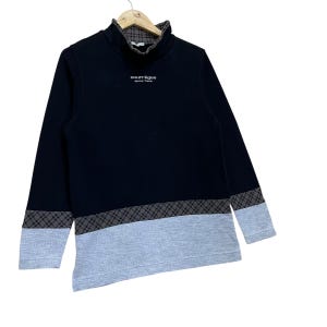 May include: Black long-sleeve sweatshirt with a high collar and gray and black plaid accents. The bottom portion of the sweatshirt and the sleeves are gray. The text "courr&egrave;ges sports futur" is printed on the chest.