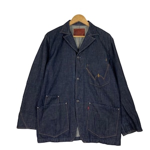 Può includere: Un blazer in denim blu scuro con revers a lancia classico e chiusura a tre bottoni. La giacca presenta due ampie tasche frontali, un taschino sul petto e una piccola etichetta rossa. Il blazer è appeso a una gruccia di legno.