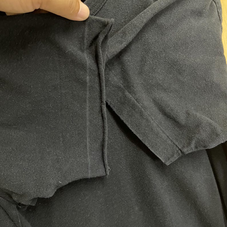 May include: Close-up of a black t-shirt, showing the sleeve and shoulder seam. The fabric appears to be a soft, slightly worn material. The image highlights the construction and texture of the garment.