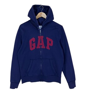Peut inclure: Sweat à capuche bleu marine zippé avec le logo "GAP" rouge sur la poitrine. Le sweat à capuche a une capuche, une poche avant et une fermeture éclair intégrale. Le vêtement est présenté sur un cintre en bois.