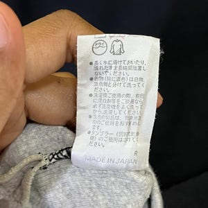 May include: A close-up of a white tag with Japanese text and symbols, including a dry cleaning symbol. The tag reads "MADE IN JAPAN" at the bottom. The tag is attached to a gray garment.