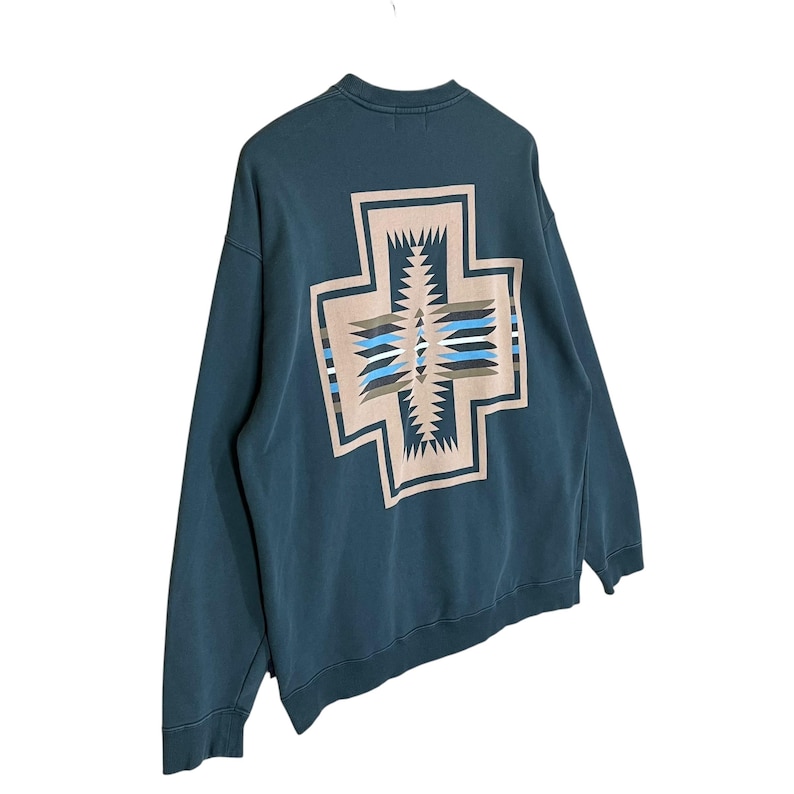 以下が含まれることがあります： 背中に大きな幾何学的な十字のデザインが施された、ティール色のスウェットシャツ。十字はベージュ、青、白の模様が特徴です。スウェットシャツは長袖でクルーネックで、カジュアルウェアに適しています。