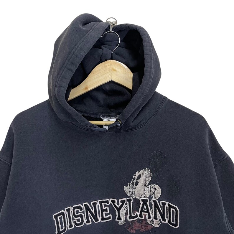 Puede incluir: Sudadera con capucha negra con un gr&aacute;fico descolorido de Mickey Mouse y el texto "DISNEYLAND" en letras blancas.