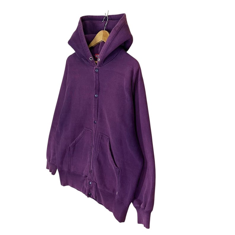 May include: A deep purple hooded sweatshirt with a button-up front and two front pockets. The sweatshirt is hanging on a wooden hanger. The fabric appears to be a soft, worn material.