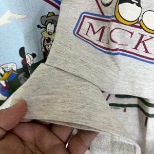 Puede incluir: Primer plano de una camiseta de manga larga gris claro con la palabra "MICK" en un dise&ntilde;o rectangular rojo y azul. La camiseta tambi&eacute;n muestra personajes de dibujos animados, incluyendo Mickey Mouse, Goofy y Donald Duck. La manga est&aacute; doblada.