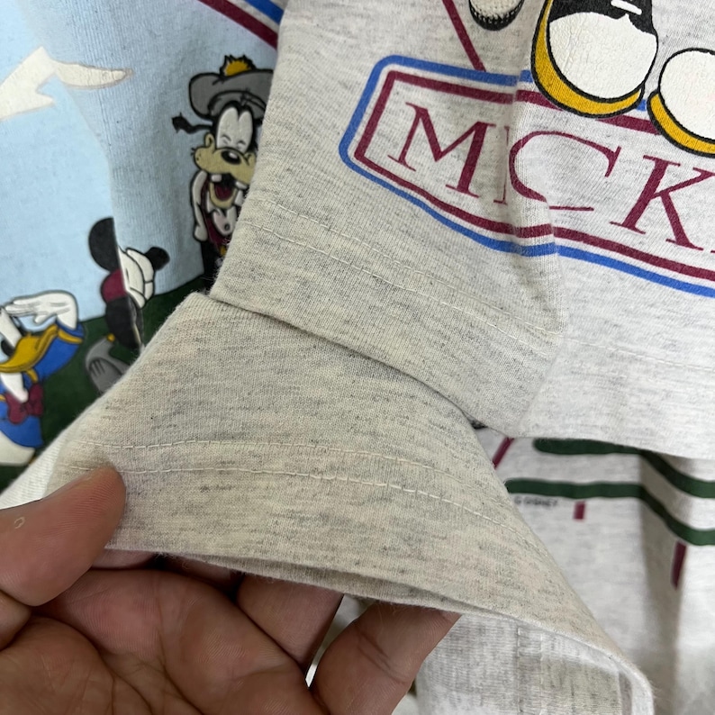 Puede incluir: Primer plano de una camiseta de manga larga gris claro con la palabra "MICK" en un dise&ntilde;o rectangular rojo y azul. La camiseta tambi&eacute;n muestra personajes de dibujos animados, incluyendo Mickey Mouse, Goofy y Donald Duck. La manga est&aacute; doblada.