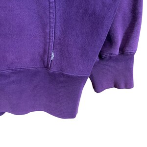 May include: Close-up of a purple sweatshirt. The ribbed cuffs and waistband are a darker shade of purple. The fabric has a soft texture, and the stitching is visible along the seams.