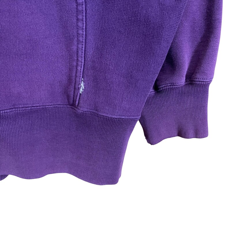 May include: Close-up of a purple sweatshirt. The ribbed cuffs and waistband are a darker shade of purple. The fabric has a soft texture, and the stitching is visible along the seams.