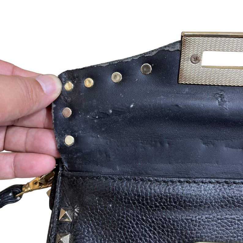 May include: Close-up of a black leather handbag with gold-colored studs and a gold-tone metal clasp. The bag shows signs of wear and tear, with some peeling of the leather. The interior is visible, revealing a textured surface.