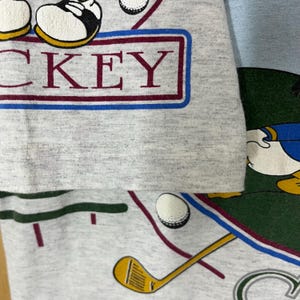 Puede incluir: Una camiseta gris con un dise&ntilde;o tem&aacute;tico de golf, que incluye la palabra "MICKEY" en un recuadro burdeos y azul. La camiseta tambi&eacute;n muestra pelotas de golf, un palo de golf y un personaje de dibujos animados.