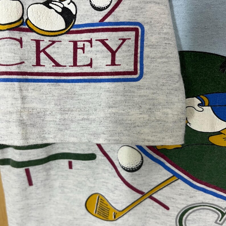 Puede incluir: Una camiseta gris con un dise&ntilde;o tem&aacute;tico de golf, que incluye la palabra "MICKEY" en un recuadro burdeos y azul. La camiseta tambi&eacute;n muestra pelotas de golf, un palo de golf y un personaje de dibujos animados.