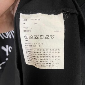 May include: A black clothing care label with white text and symbols. The label includes the brand name "Commes des Garcons", the product number "PE-T066", the size "L", and the year "AD2018". The label also includes care instructions in Japanese.