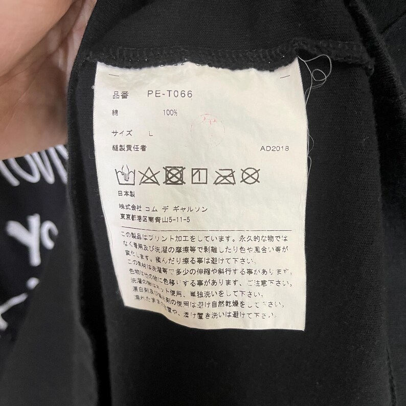 May include: A black clothing care label with white text and symbols. The label includes the brand name "Commes des Garcons", the product number "PE-T066", the size "L", and the year "AD2018". The label also includes care instructions in Japanese.