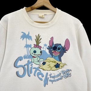 K&ouml;nnte beinhalten: Wei&szlig;er Pullover mit einem blauen und hellblauen Aufdruck von Stitch und Angel aus Lilo & Stitch. Der Aufdruck zeigt Stitch und Angel an einem Strand mit Palmen. Der Text "Stitch Tropical Terror Experiment 626" ist unter dem Aufdruck gedruckt.
