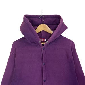 May include: A purple hooded sweatshirt with a button-up front. The sweatshirt is hanging on a wooden hanger. The brand name "Dickies" is visible on a small tag near the top button.