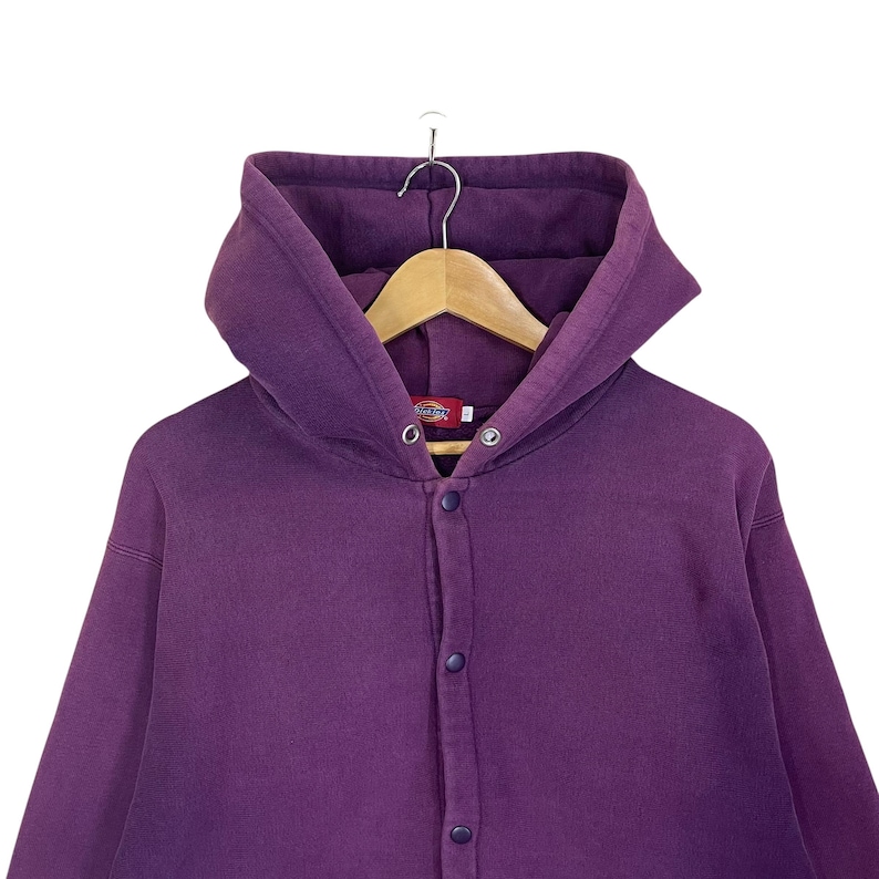 May include: A purple hooded sweatshirt with a button-up front. The sweatshirt is hanging on a wooden hanger. The brand name "Dickies" is visible on a small tag near the top button.