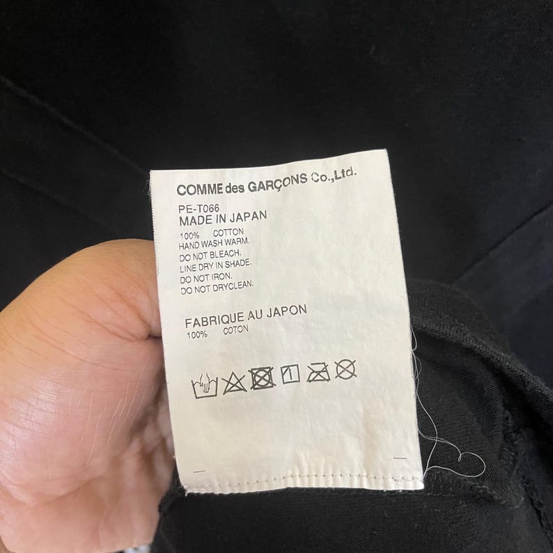 May include: White care label with black text that reads "COMME des GAR&Ccedil;ONS Co.,Ltd. PE-T066 MADE IN JAPAN 100% COTTON HAND WASH WARM. DO NOT BLEACH LINE DRY IN SHADE. DO NOT IRON DO NOT DRYCLEAN. FABRIQUE AU JAPON 100% COTON" and a series of black and white care symbols.