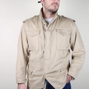 Könnte beinhalten: Beige Jacke im Militärstil mit vier Vordertaschen, Knopfverschluss und hohem Kragen. Die Jacke hat Schulterklappen und wird mit einer marineblauen Baseballkappe und Jeans getragen. Ein lässiger Vintage-Stil.