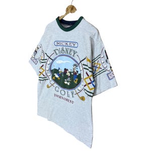 Puede incluir: Camiseta vintage gris claro con un dise&ntilde;o de Mickey Mouse Disney Golf Tournament. La camiseta tiene un cuello verde y azul marino y un gr&aacute;fico de Mickey, Donald y Goofy jugando al golf. Las mangas tienen patrones repetidos con tem&aacute;tica de golf.