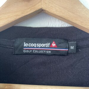 May include: Black golf shirt tag with white text that reads "le coq sportif GOLF COLLECTION" and a red rooster logo. The tag also has a white stripe with a red and blue stripe above it. The size "M" is printed in white on a black tag.