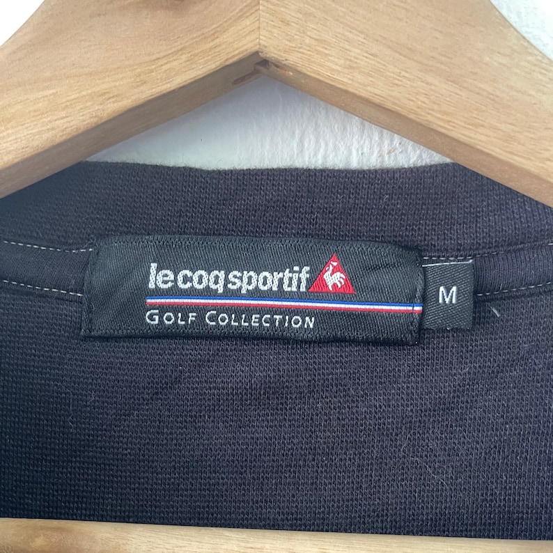 May include: Black golf shirt tag with white text that reads "le coq sportif GOLF COLLECTION" and a red rooster logo. The tag also has a white stripe with a red and blue stripe above it. The size "M" is printed in white on a black tag.
