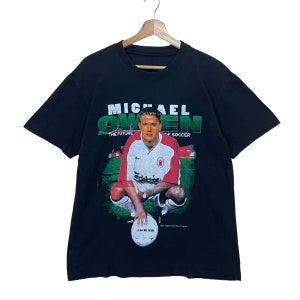 May include: Black t-shirt with a graphic print of Michael Owen, a soccer player, in a red and white jersey. The text "Michael Owen" is printed in white above the image, and the text "The Future of Soccer" is printed in white below the image.