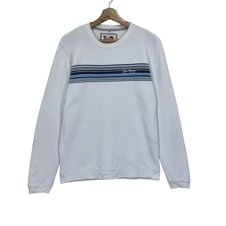 Pu&ograve; includere: Felpa bianca a maniche lunghe con una fascia blu e grigia a righe sul petto. Il testo "Surf Style" &egrave; stampato sulla fascia.
