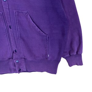 May include: A purple button-up sweatshirt with two front pockets and a ribbed hem and cuffs. The sweatshirt has a relaxed fit and a vintage look. The buttons are a matching purple color. The fabric appears to be a soft, comfortable material.