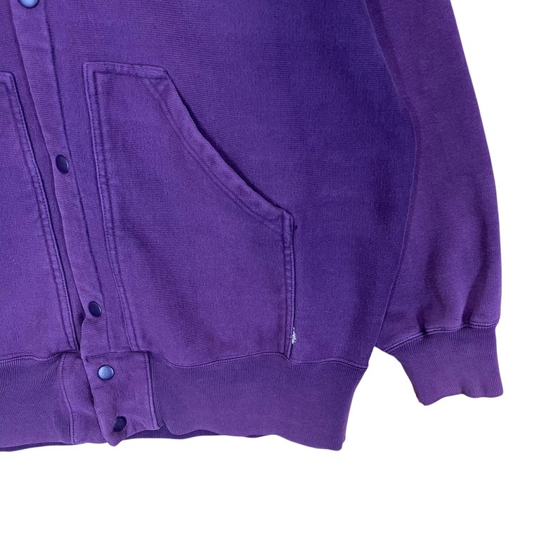 May include: A purple button-up sweatshirt with two front pockets and a ribbed hem and cuffs. The sweatshirt has a relaxed fit and a vintage look. The buttons are a matching purple color. The fabric appears to be a soft, comfortable material.