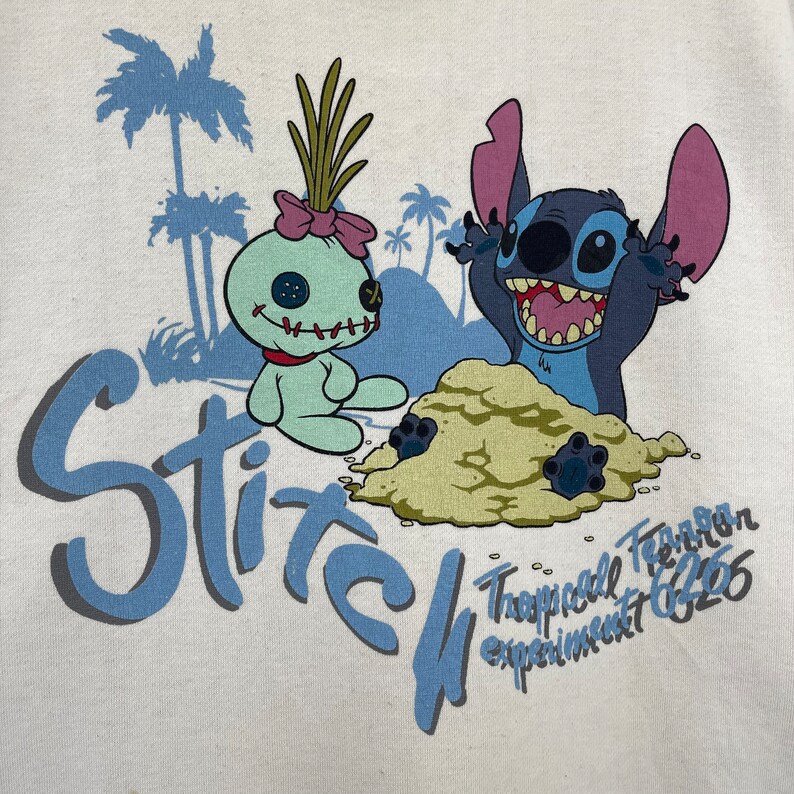 K&ouml;nnte beinhalten: Ein wei&szlig;es T-Shirt mit einem blauen und gr&uuml;nen Motiv von Stitch und Angel aus Lilo & Stitch. Das Motiv zeigt Stitch und Angel an einem Strand mit Palmen. Der Text "Stitch Tropical Experiment 626" ist unter dem Motiv gedruckt.