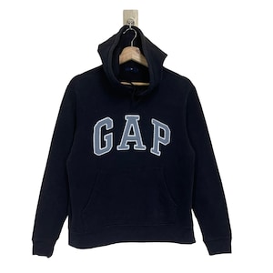 Może przedstawiać: Czarna bluza z kapturem z napisem "GAP" w dużych, szarych literach na piersi. Bluza ma przednią kieszeń i kaptur ze sznurkiem. Logo GAP jest obrysowane na biało.