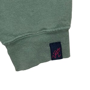 Pu&ograve; includere: Primo piano di una manica di felpa verde salvia. Il polsino presenta un'etichetta blu navy con una grafica rossa. Il tessuto sembra essere un materiale a maglia morbido. Sfondo bianco.