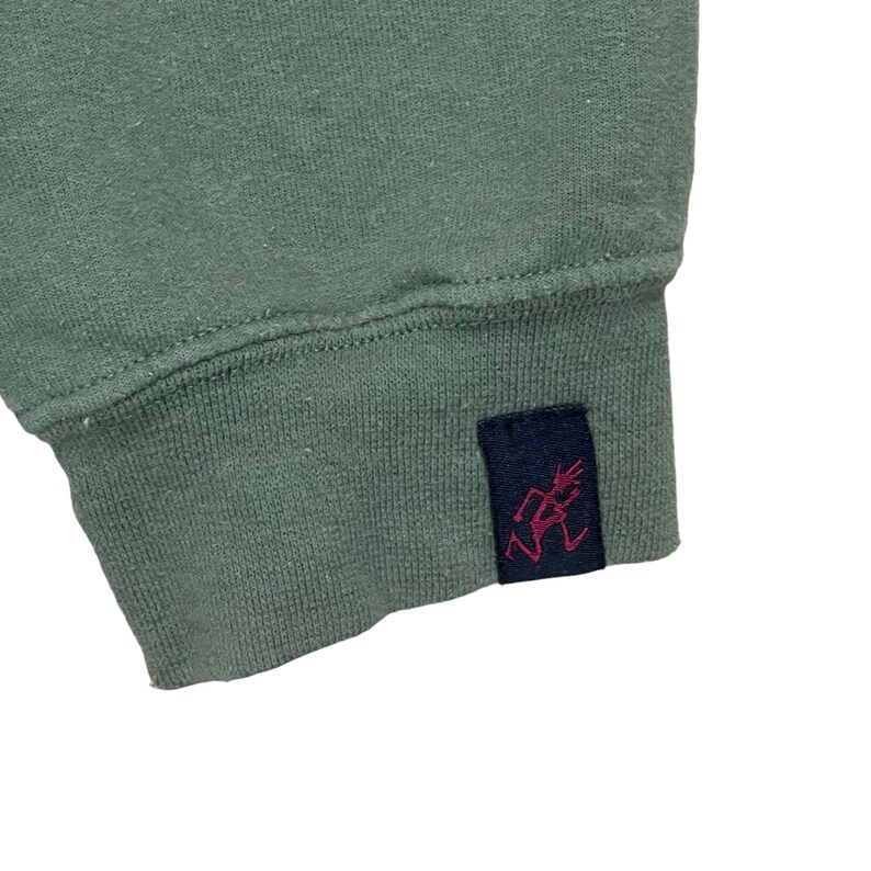 Pu&ograve; includere: Primo piano di una manica di felpa verde salvia. Il polsino presenta un'etichetta blu navy con una grafica rossa. Il tessuto sembra essere un materiale a maglia morbido. Sfondo bianco.