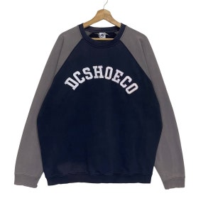 May include: A navy blue and gray DC SHOECO sweatshirt. The sweatshirt has a navy blue body with gray sleeves. The text "DCSHOECO" is arched across the chest in white lettering. The sweatshirt is hanging on a wooden hanger.