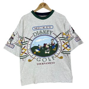 Puede incluir: Camiseta gris claro con un dise&ntilde;o de Mickey Mouse Disney Golf Tournament. La camiseta tiene cuello y mangas verdes con personajes de dibujos animados jugando al golf. El texto "Mickey Disney Golf Tournament" est&aacute; impreso en la parte delantera.