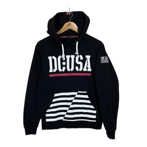 May include: Black hooded sweatshirt with the text "DC USA" in white block letters, a red stripe, and a pocket with white and black stripes. A small American flag patch is on the right sleeve.