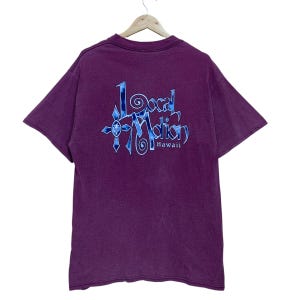 May include: A purple t-shirt featuring the text "Local Motion Hawaii" in a blue and white design on the back. The shirt is hanging on a wooden hanger. The t-shirt has short sleeves and is against a white background.