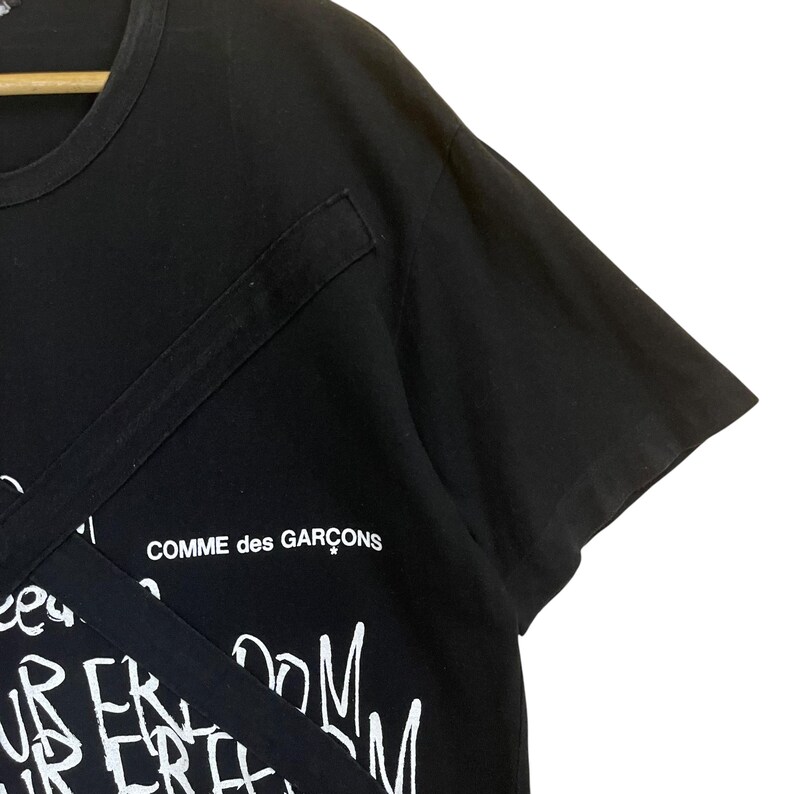 May include: Black t-shirt with a white graphic print that says "Comme des Gar&ccedil;ons" and has a white graphic print of a design with the words "freedom" and "room" repeated.