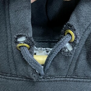 Puede incluir: Primer plano de una sudadera con capucha negra con cord&oacute;n. El cord&oacute;n tiene dos ojales met&aacute;licos y est&aacute; deshilachado en los extremos.