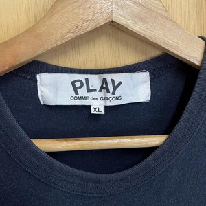 May include: Black crew neck t-shirt with a white label that reads "PLAY COMME des GAR&Ccedil;ONS" and "XL". The shirt is hanging on a wooden hanger.