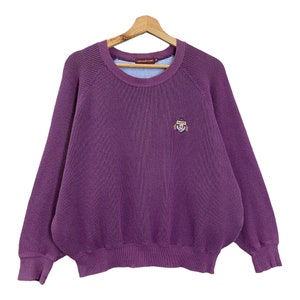 May include: A purple ribbed crew neck sweater with long sleeves. The sweater features a small embroidered emblem on the chest and a light blue neckline. The sweater is suspended on a wooden hanger.
