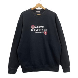 Puede incluir: Sudadera negra con texto blanco y rojo "ZYXON GOLFSTYLE Munsingwear". La sudadera está colgada de una percha de madera. El logotipo de ZYXON es un círculo rojo.
