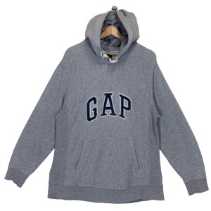 Könnte beinhalten: Grauer Kapuzenpullover mit dem Wort "GAP" in marineblauen Buchstaben auf der Brust. Der Pullover hat eine Vordertasche und eine Kapuze mit Kordelzug. Das Kleidungsstück wird auf einem Holzkleiderbügel präsentiert.