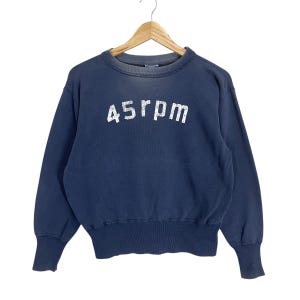 Könnte beinhalten: Ein marineblaues Sweatshirt mit dem weißen Text "45rpm". Der Vintage-Rundhals-Pullover hat lange Ärmel und gerippte Bündchen und Saum. Das Sweatshirt hängt an einem Holzkleiderbügel.